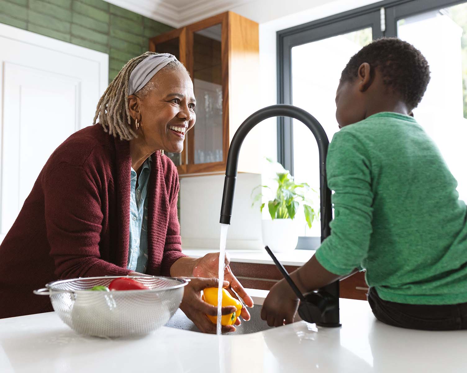 A smiling elderly woman and a boy wash vegetables in a sunny kitchen