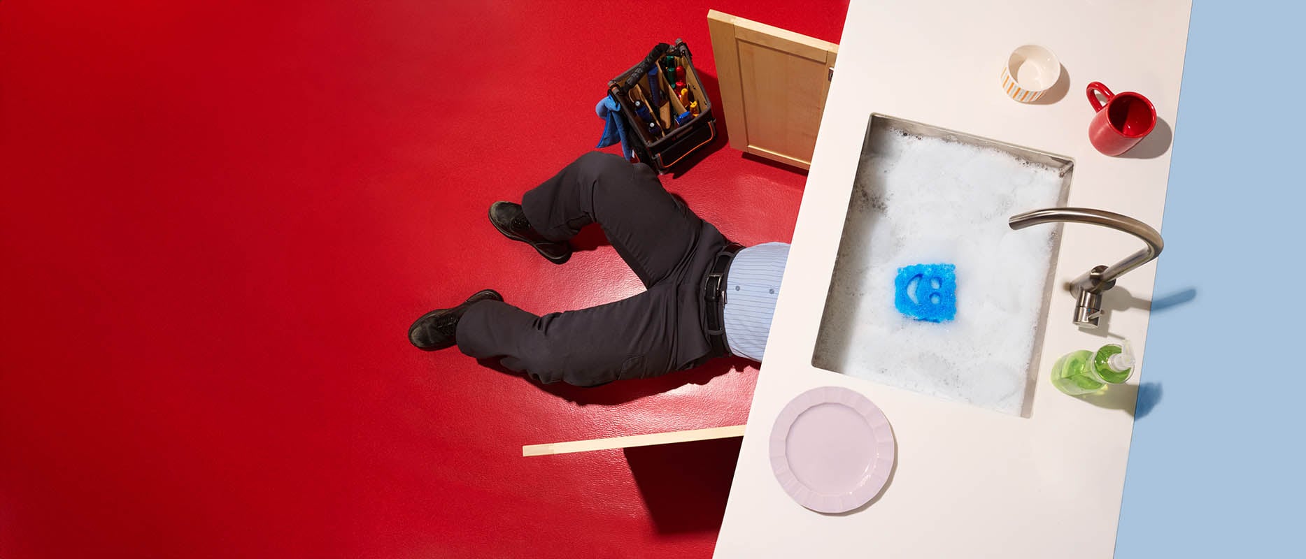 Overhead shot of a sink on the right, and red floor, and a workers legs coming out from under the sink.