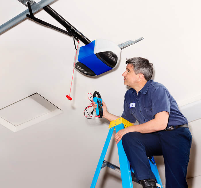 Garage door technician on a ladder inspecting an automatic garage door opener with a handheld electrical testing tool.